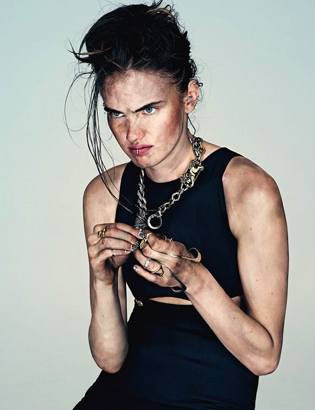 Portrait of a woman in a black dress with heavy jewelry, her face showing smudges, against a plain backdrop in a studio setting.