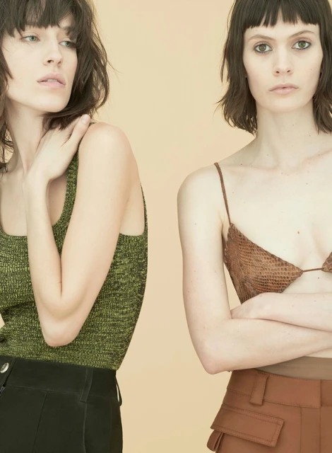 Two adult women pose indoors against a solid background, wearing fashionable tops and high-waisted trousers in a studio shot.