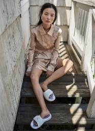Woman in a shiny beige blouse and skirt sitting on weathered outdoor wooden stairs wearing white slides
