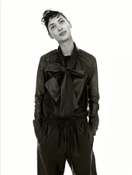 Portrait of a woman wearing a dark bow-collared shirt and trousers with hands in pockets against a plain background