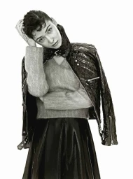 Model with short dark hair wearing a textured grey sweater and black quilted jacket against a white background.