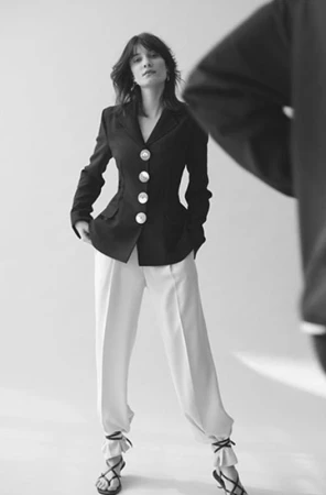 Black and white portrait of a woman wearing a dark blazer with large buttons and light trousers against a seamless backdrop
