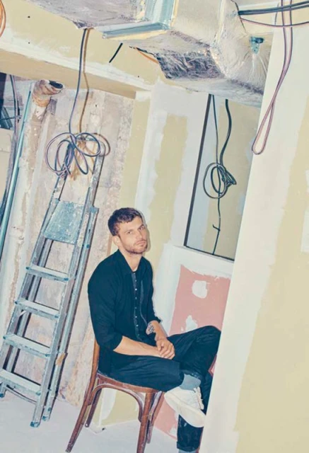 Adult man sits on a wooden chair in an unfinished room with a ladder, exposed wires, and unpainted walls in a neutral indoor setting.
