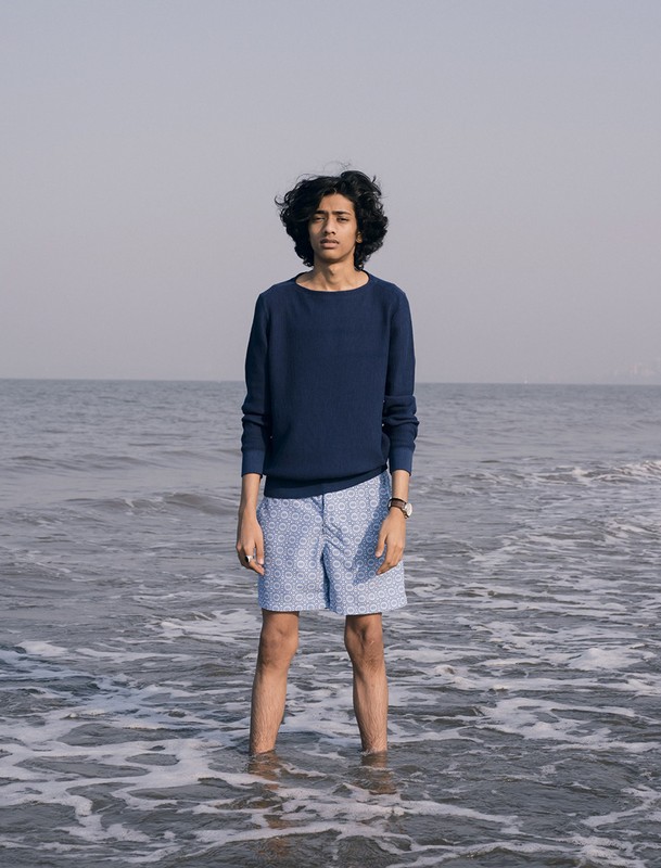 Young man standing ankle-deep in the ocean wearing a navy sweater and patterned light blue shorts styled by Dan May.
