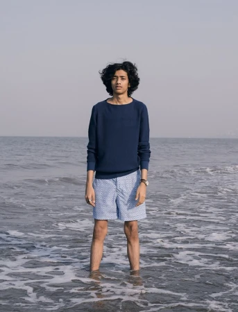 Young man standing ankle-deep in the ocean wearing a navy sweater and patterned light blue shorts styled by Dan May.