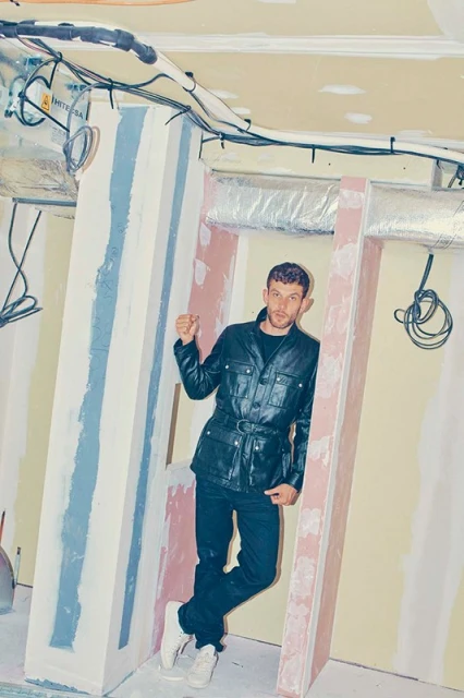 Man in black jacket and jeans poses indoors in a construction-like setting with exposed wires and wall framework.