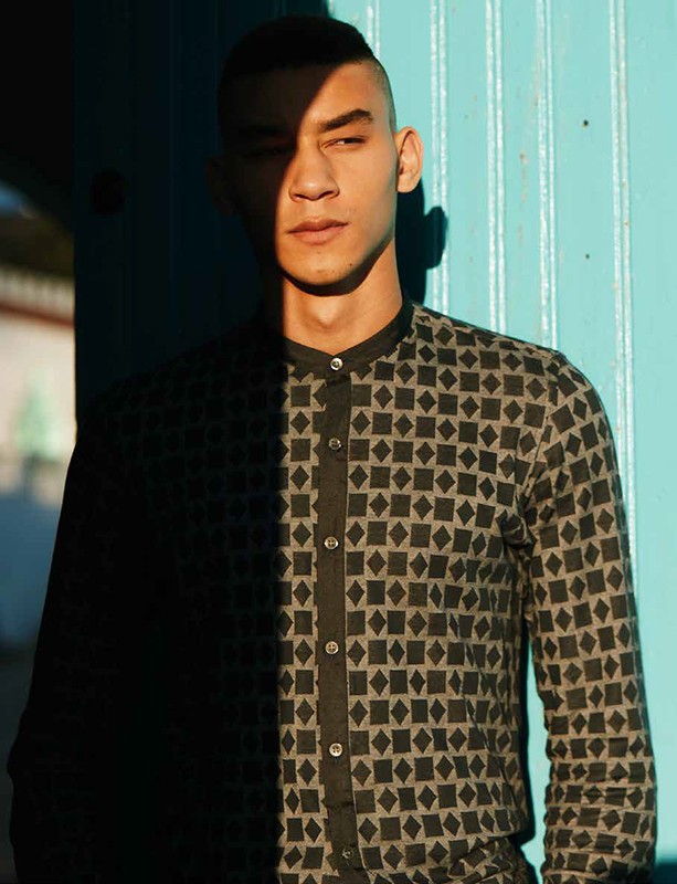 Portrait of an adult male in a patterned shirt standing against a teal wall with sunlight creating shadows on his face and body.