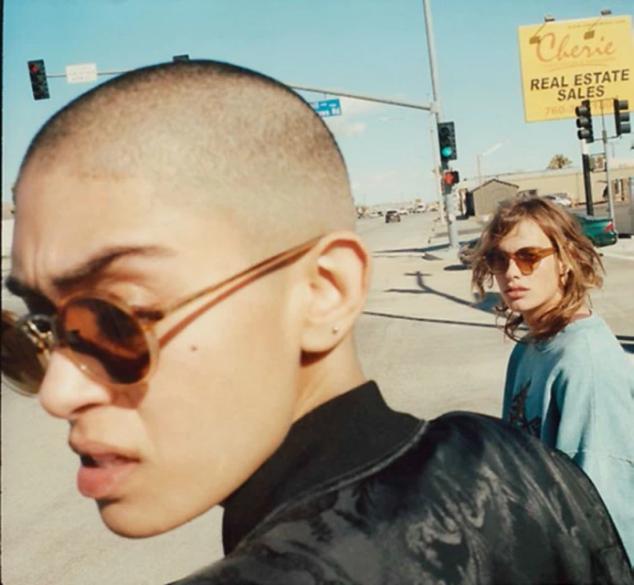 Two stylish people in sunglasses on a sunny city street. One person with a shaved head is in the foreground and another behind.