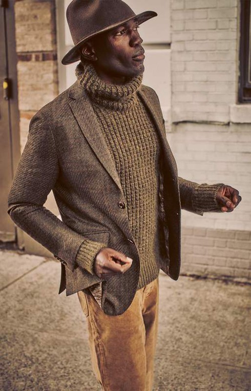 An African American man stands on a sidewalk with a dark brown hat, olive green sweater, and brown blazer in an urban setting.