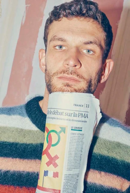Portrait of a man holding a newspaper with gender symbols against a painted backdrop, showcasing a blend of contemporary editorial and personal style.