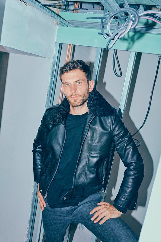 Indoor portrait of adult male in leather jacket posing in front of an unfinished wall with exposed wiring and structural supports.