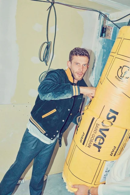 Man holding insulation material in an indoor construction setting, posing with an inquisitive look and wearing casual jacket and pants.
