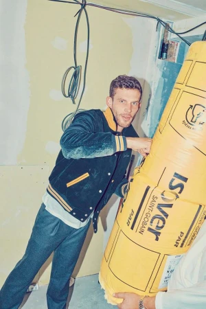 Man holding insulation material in an indoor construction setting, posing with an inquisitive look and wearing casual jacket and pants.