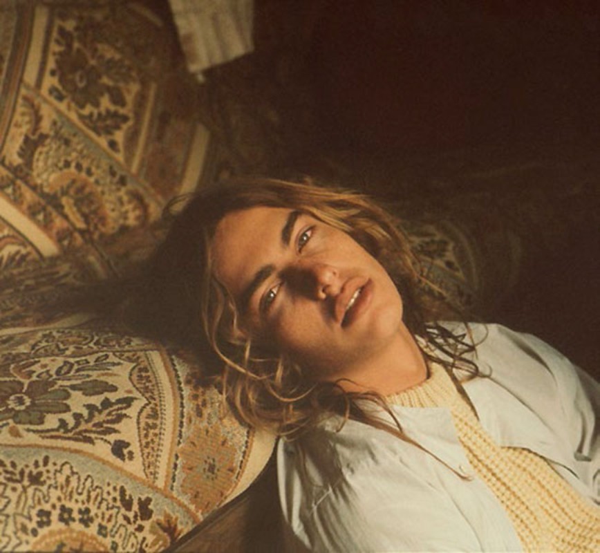 A person with long brown hair is lying on a patterned cushion, gazing directly at the viewer with a neutral facial expression.