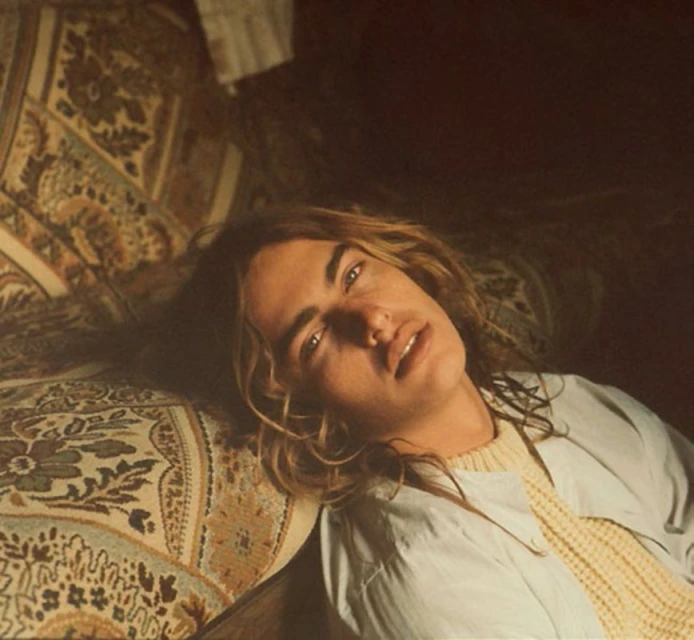 A person with long brown hair is lying on a patterned cushion, gazing directly at the viewer with a neutral facial expression.
