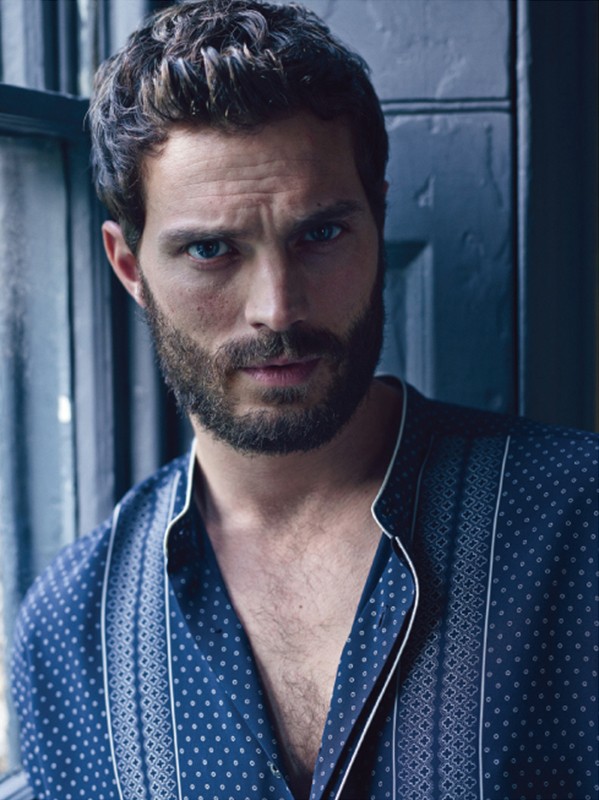 Portrait of a bearded man with dark hair, wearing a patterned blue shirt, standing by a window with natural light.