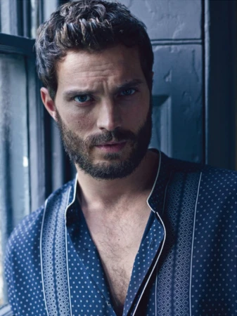 Portrait of a bearded man with dark hair, wearing a patterned blue shirt, standing by a window with natural light.