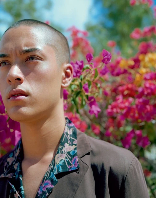 A young adult male with a shaved head poses in a close-up shot, wearing a brown shirt with a floral-print collar, in a garden with colorful flowers.