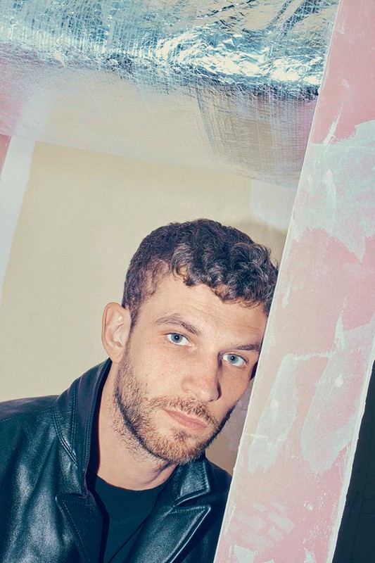 Man in a black jacket leans against a pink surface with a silver ceiling visible. Portrait-style photo with cool color grading and texture.
