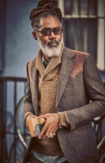 Stylish mature man with dreadlocks, beard, and glasses wearing a tweed blazer and sweater, holding a box in an urban setting.