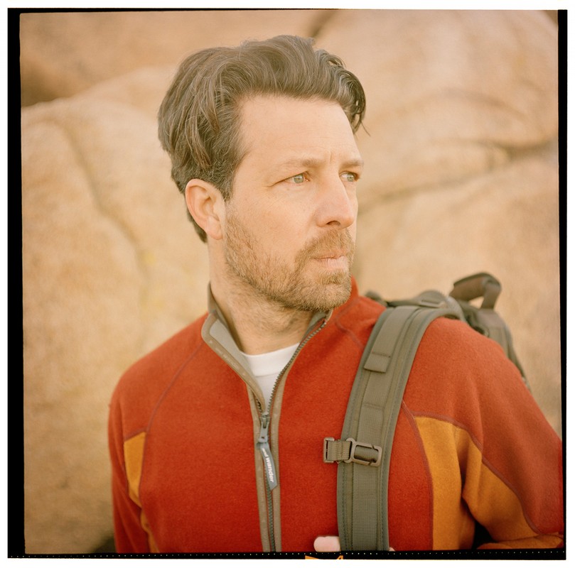 Outdoor portrait of a man with a backpack in front of rock formations, wearing a rust-colored fleece jacket and looking away.