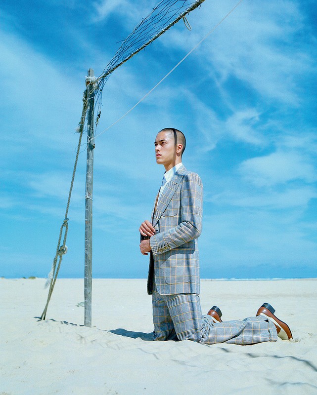 An adult male kneeling on a white sand beach near a volleyball net wearing a suit under a sunny sky. The man is looking left.