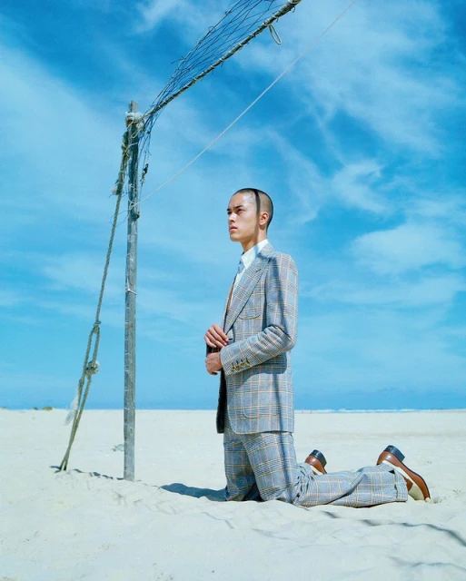An adult male kneeling on a white sand beach near a volleyball net wearing a suit under a sunny sky. The man is looking left.