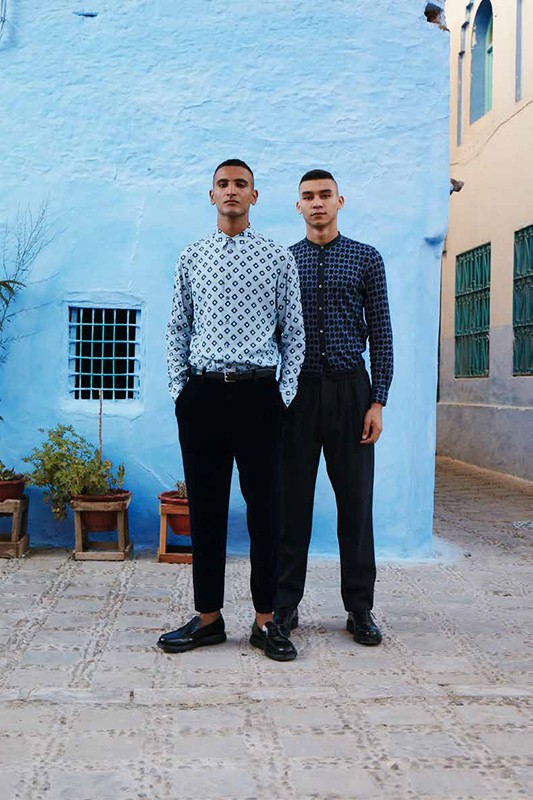 Two adult males posing outdoors against a blue wall in patterned shirts and black pants, standing on stone-paved ground with potted plants.