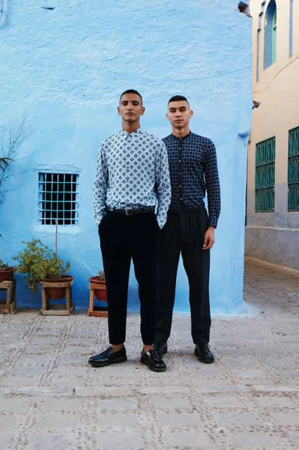 Two adult males posing outdoors against a blue wall in patterned shirts and black pants, standing on stone-paved ground with potted plants.