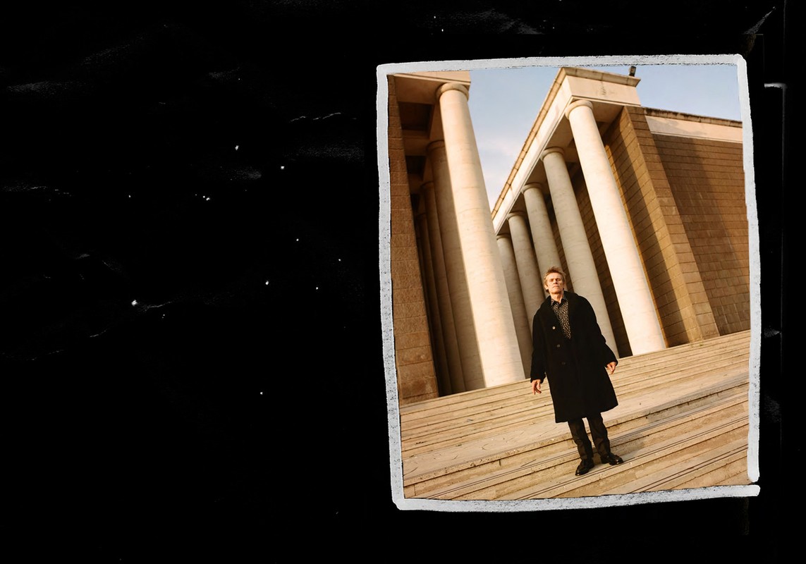 Man in coat stands on stairs in front of columned building on black background in fashion shot with editorial style and neutral palette.