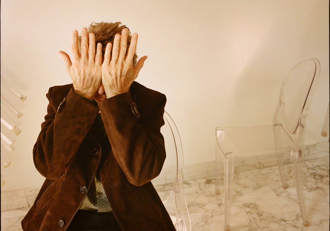 Older adult with their hands covering their face, seated on a transparent chair in an indoor setting with a neutral backdrop.