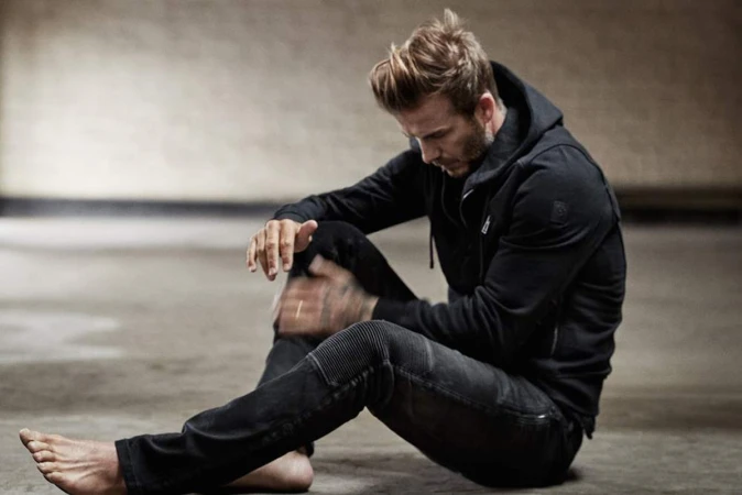 David Beckham seated on the floor wearing a black hoodie and dark trousers with bare feet in a loft space