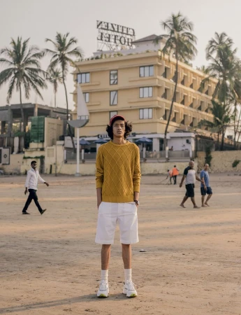 Young man in yellow knit sweater and white shorts standing on a sandy beach with a hotel in the background.