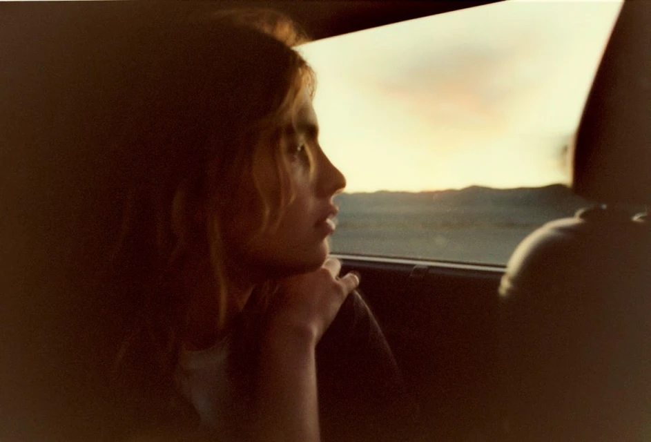 Teenager looks out car window at sunset over desert landscape. Soft focus and warm tones evoke a sense of nostalgia and reflection.