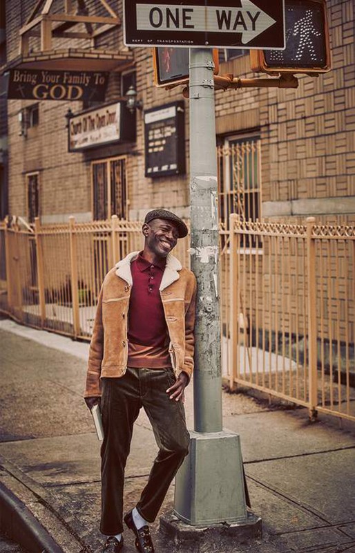 Smiling man in a jacket and cap stands on a sidewalk next to a street sign in front of a brick building in an urban environment.