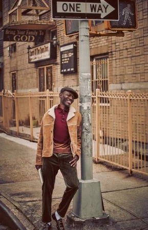 Smiling man in a jacket and cap stands on a sidewalk next to a street sign in front of a brick building in an urban environment.