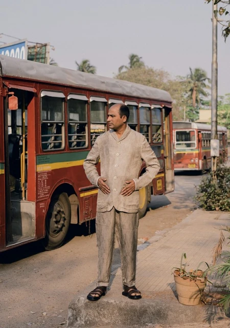 Man in beige suit stands near red bus. Outdoor shot features neutral color palette, natural lighting, and urban street scene.