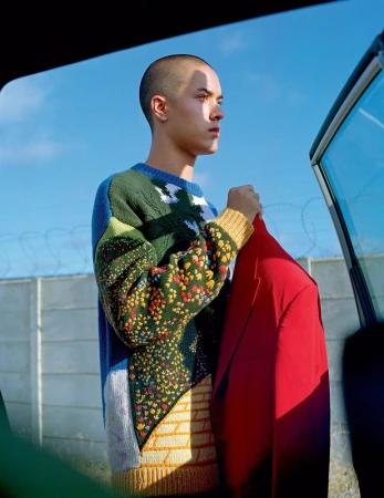 A young man with a shaved head wearing a patterned knit sweater and holding a red jacket outdoors in Los Angeles.