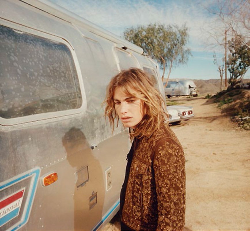 A young adult stands next to an Airstream trailer on a sunny day, wearing a brown jacket with floral patterns.