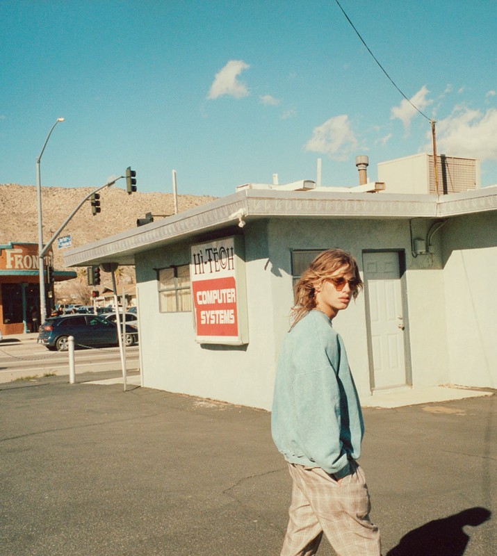 A blond man in sunglasses walks past a building with a 'Hi-Tech Computer Systems' sign on a sunny day in a desert-adjacent town.