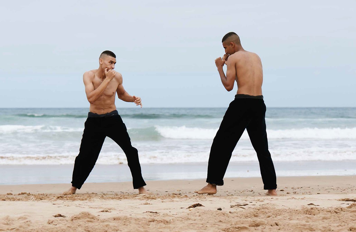 Two adult males practice martial arts on a sandy beach with waves in the background, showcasing fitness and athleticism in an outdoor setting.