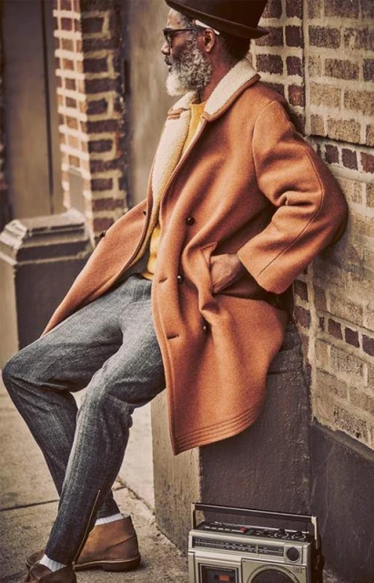 Stylish older man with gray beard wearing a coat and hat, leaning against a brick wall with a retro boombox nearby in an urban setting.