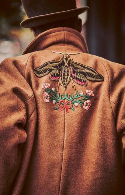 Back view of a person wearing a brown coat with a moth and floral embroidery, complemented by a brown top hat.