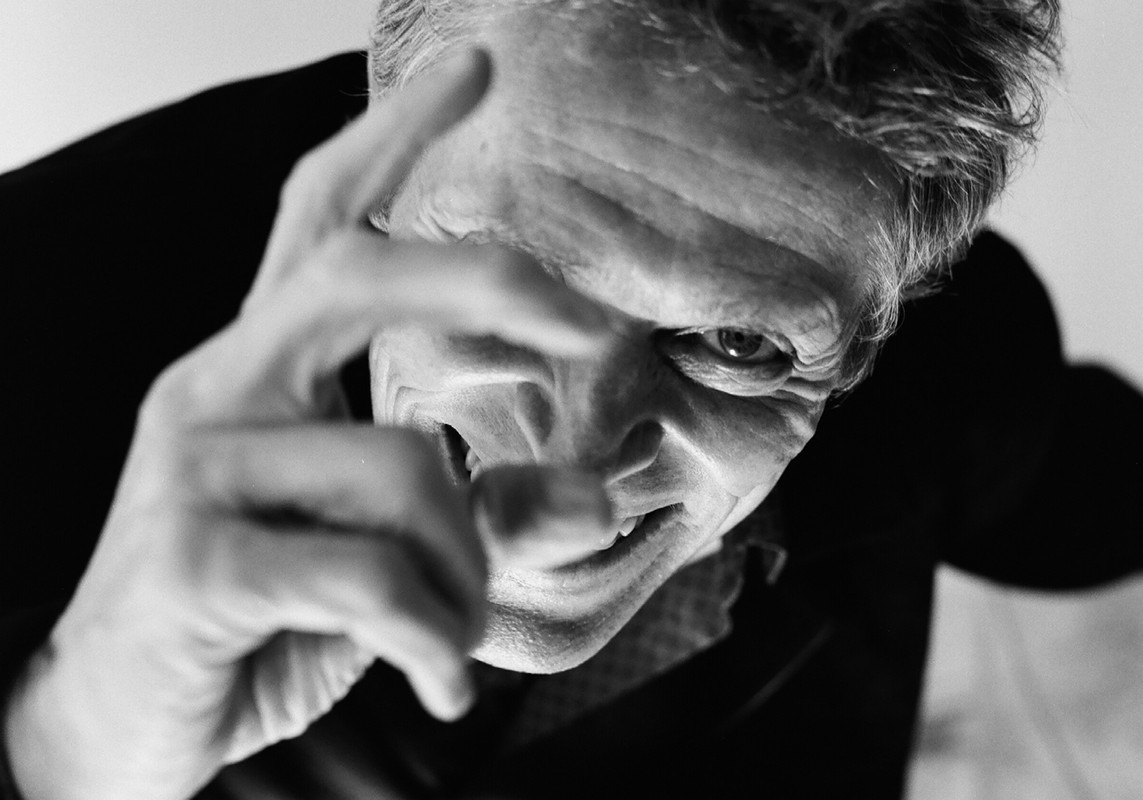 Black and white portrait of an adult man with gray hair, looking down with a slightly furrowed brow and raised hand, indoors.