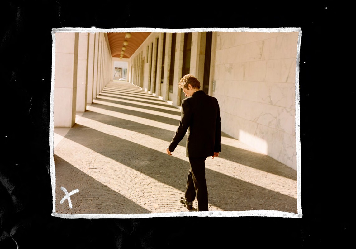 Adult male in a black suit walks along a colonnade, casting long shadows on the paving stones in an outdoor architectural space.