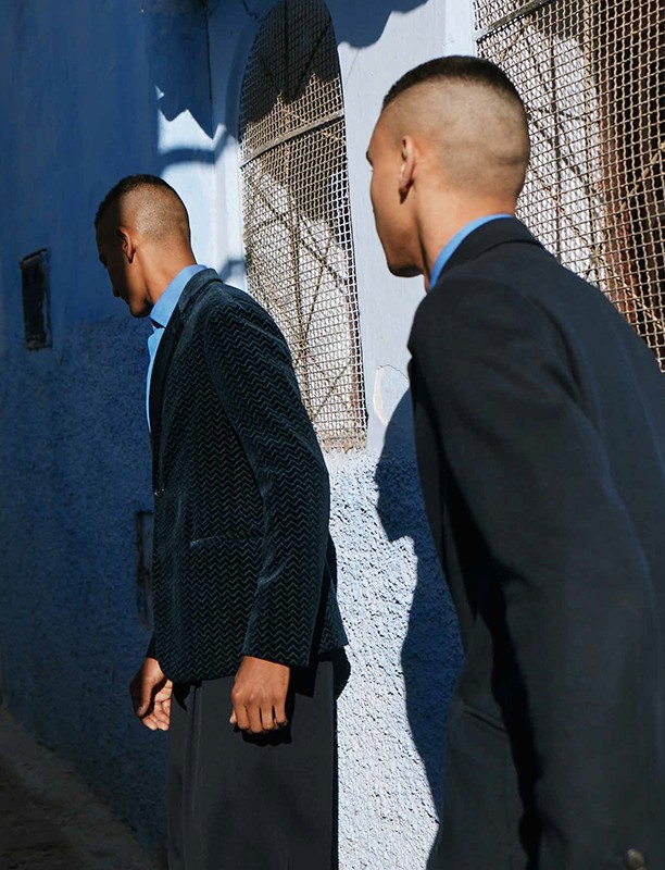 Two adult men in suits standing on a blue street. The first man wears a velvet jacket and the other a black jacket in natural light.