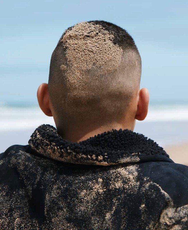 Person wearing a black jacket stands on a sandy beach covered in sand. The sea and the sky are visible in the background.