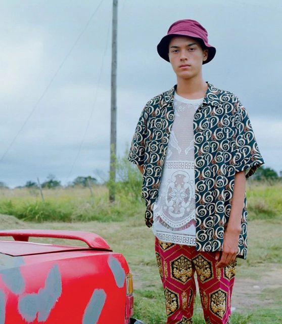 Young man in patterned shirt and pants poses next to a vintage red car in a rural landscape with a cloudy sky.