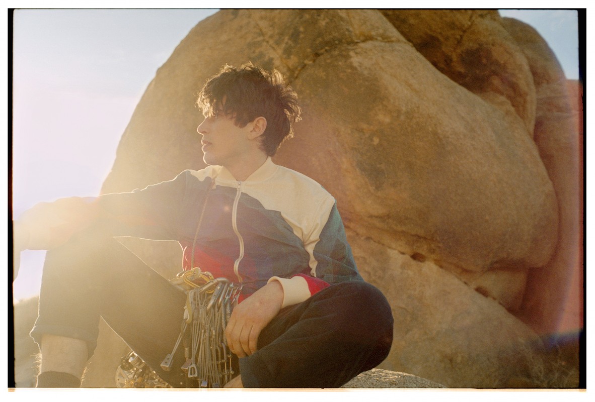 Man sits in the sun near a large rock formation, wearing a jacket and climbing gear, looking towards the left with a neutral expression.