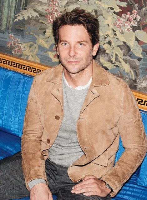 Attractive adult male with brown hair, wearing a suede jacket and seated on a blue patterned sofa in front of a floral wallpaper backdrop.